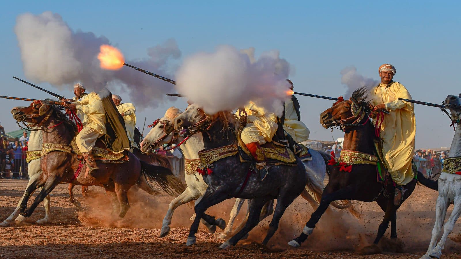 Tbourida riders in yellow robes galloping and firing their muskets in a cloud of smoke