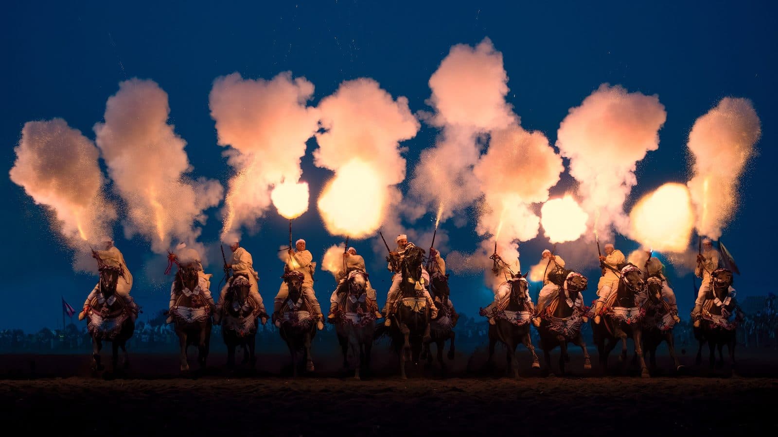 A line of tbourida riders firing their muskets at dusk, the gunpowder flashes glowing above the horses