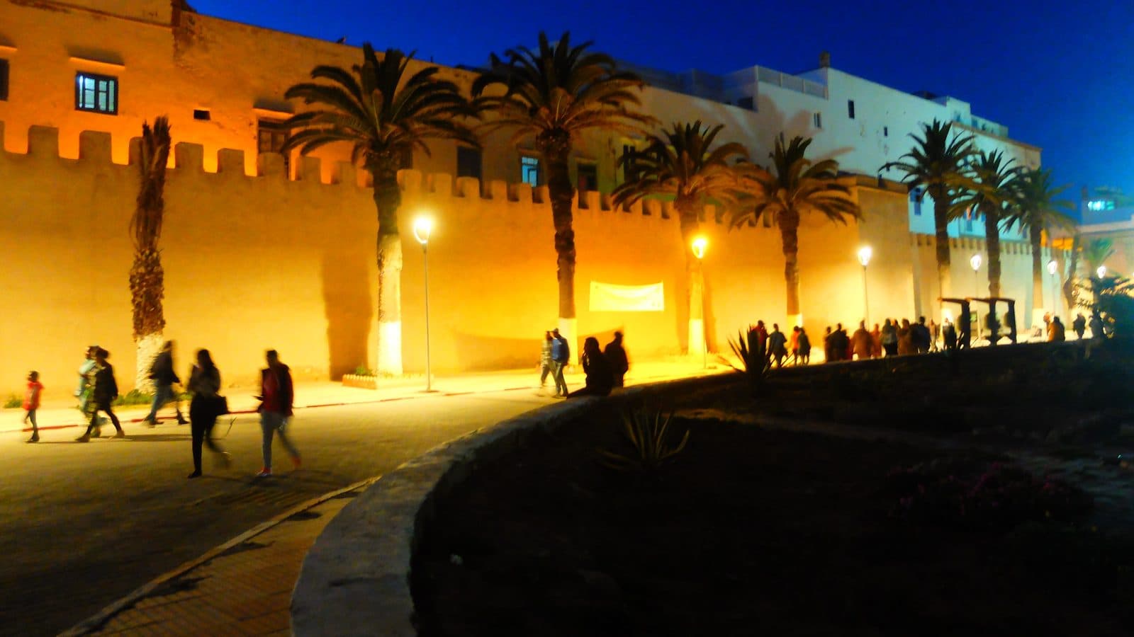 People walking along the lit-up ramparts and palm trees of Essaouira at night