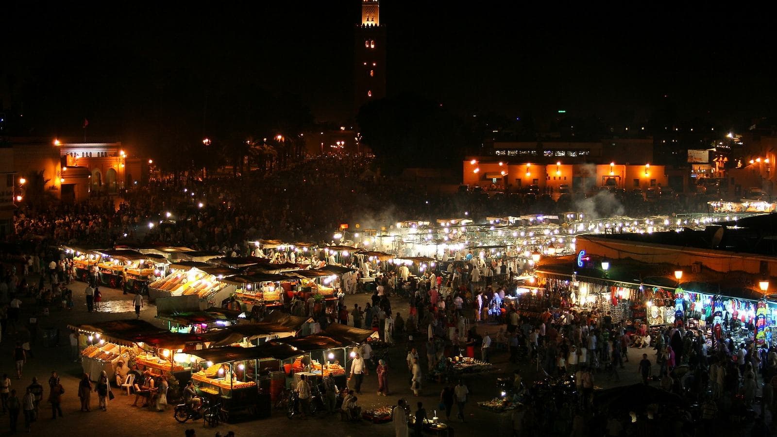 La place Jemaa el-Fna à Marrakech de nuit, pleine de stands et de monde, avec le minaret de la Koutoubia