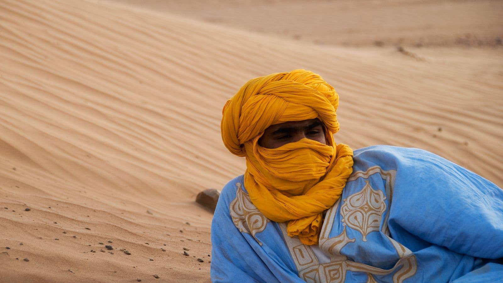 A man wearing a blue robe and a yellow turban wrapped around his face, resting on a sand dune