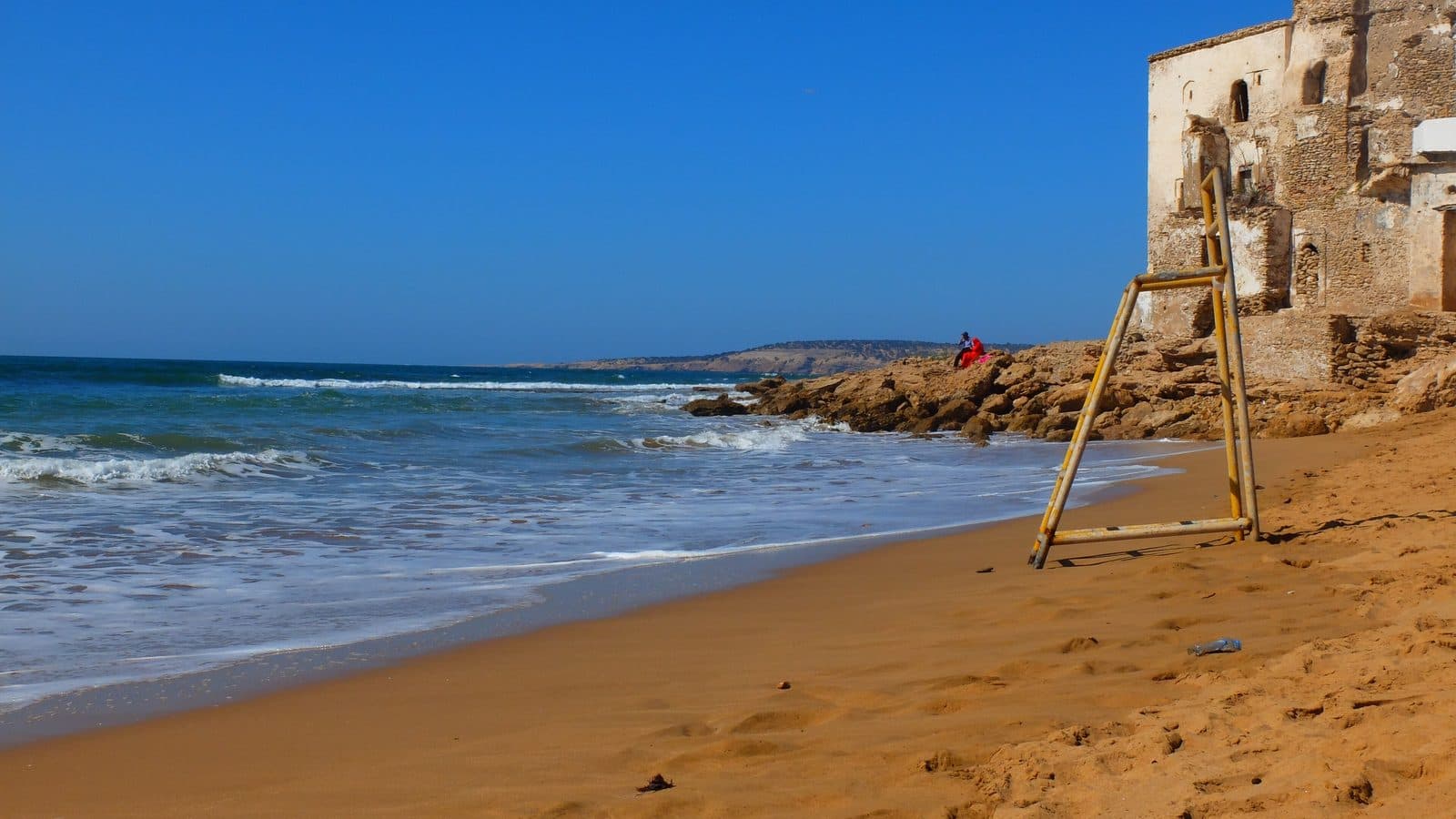 La plage dorée de Sidi Kaouki et ses vagues, avec un vieux bâtiment au bord du sable