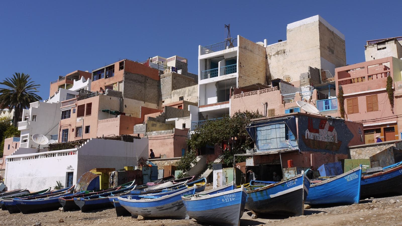 Des barques de pêche bleues tirées sur la plage sous les maisons colorées de Taghazout