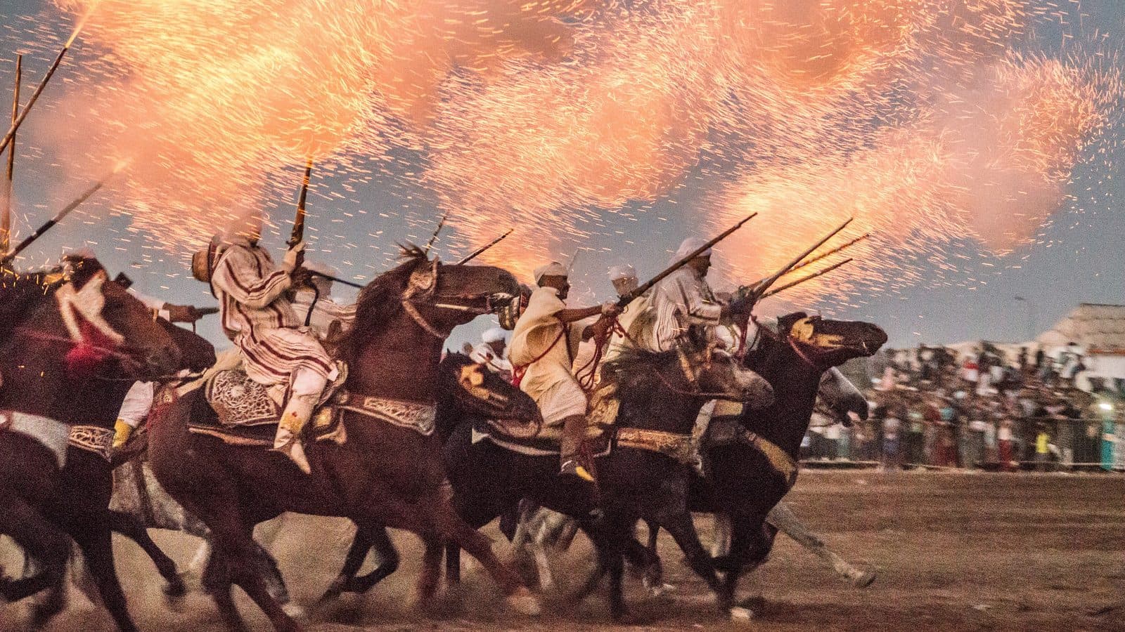 Tbourida riders galloping under a sky lit by the sparks and smoke of their muskets
