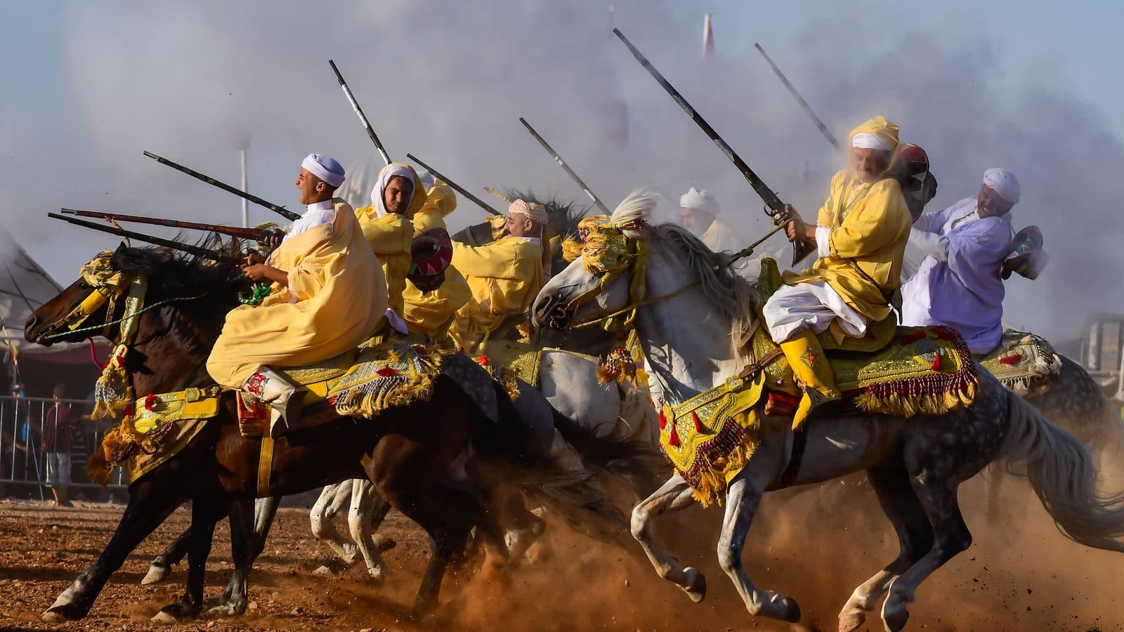 Tbourida riders in yellow robes galloping and firing their muskets on a dusty track