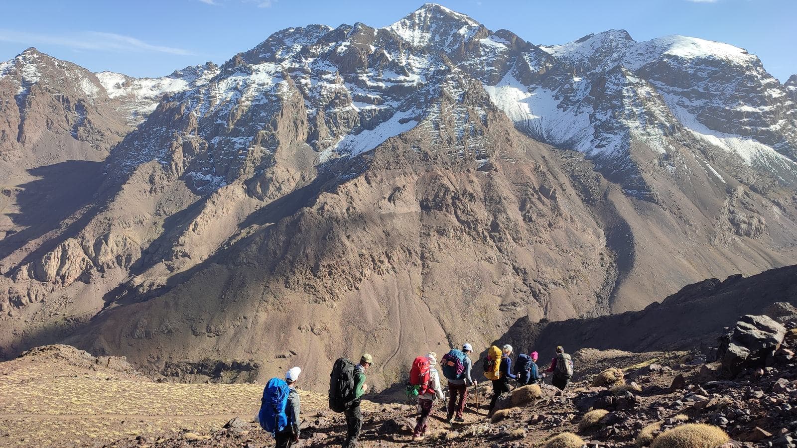 A group of hikers on a trail below the snow-streaked peaks of the Toubkal massif in the High Atlas