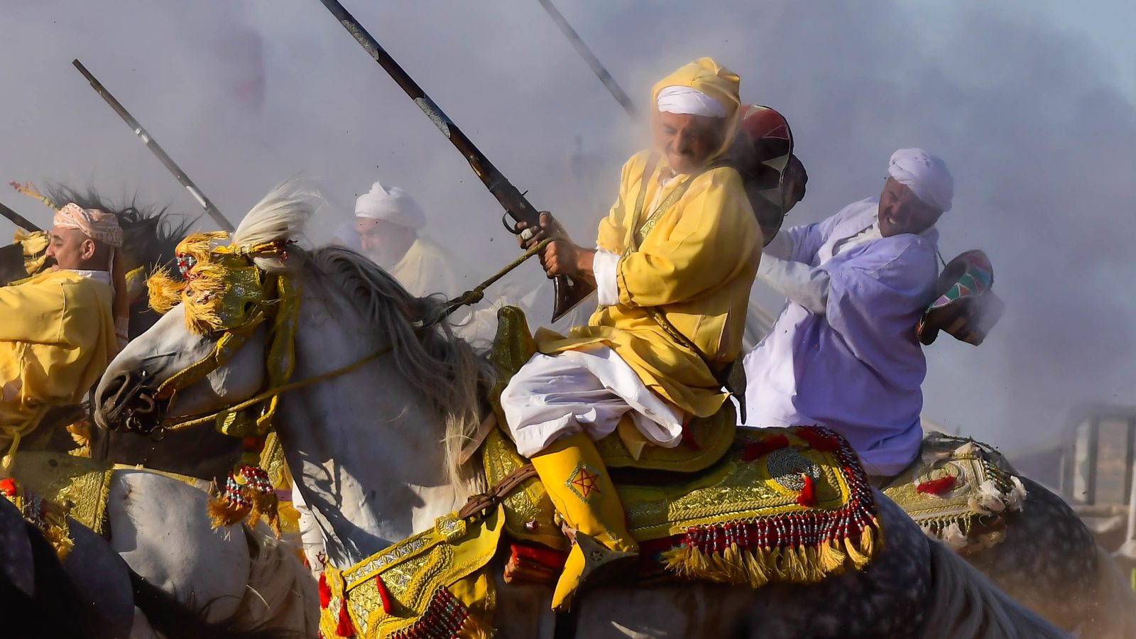 A tbourida rider in a yellow robe on a grey horse with an embroidered saddle, in a cloud of dust