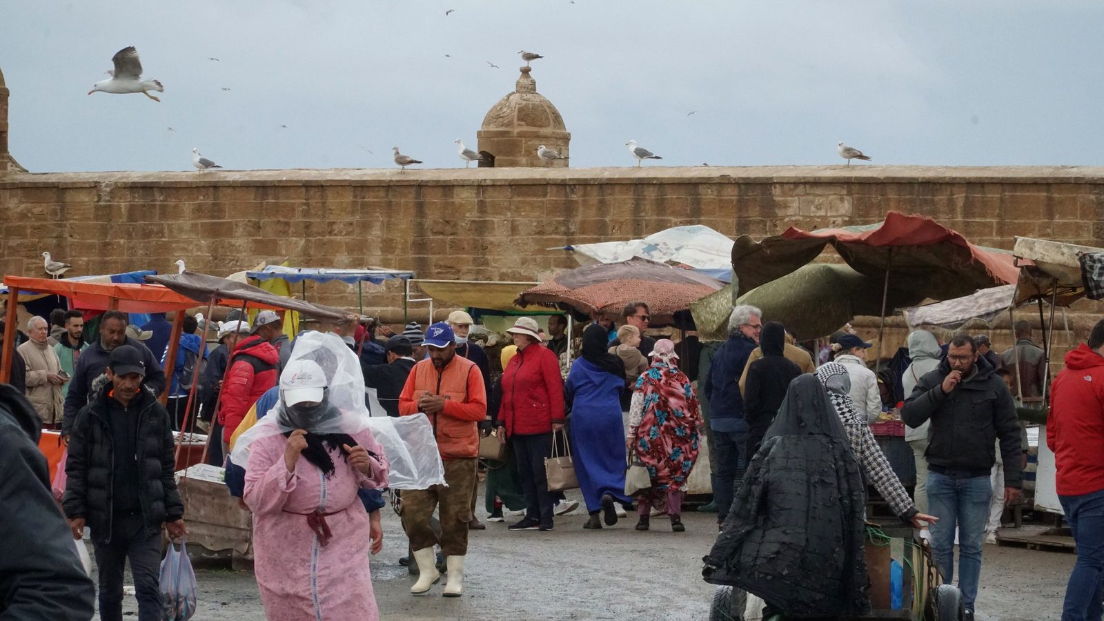 Passants en manteaux et vestes au marché le long des murs du port d'Essaouira par un jour nuageux
