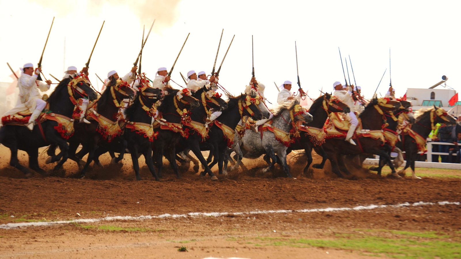 Riders in white robes and turbans charging in a line with raised rifles at a horse festival in El Jadida