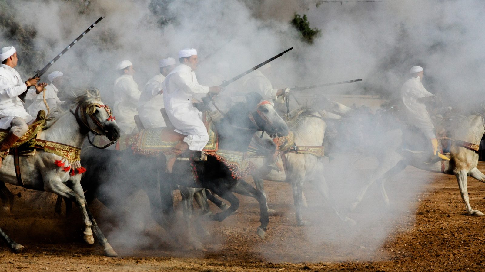 Riders in white galloping through thick gunpowder smoke on a dusty track