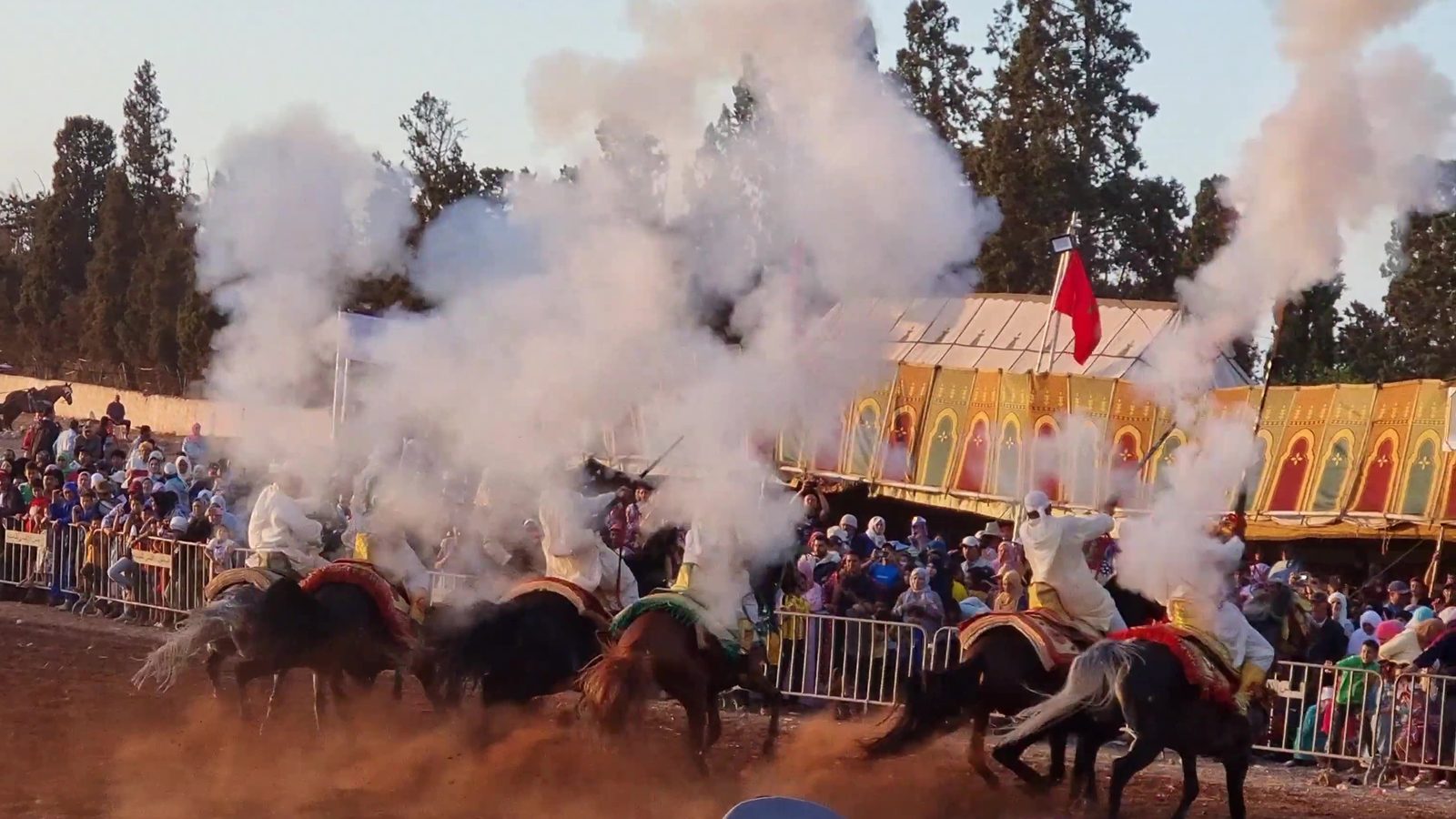 Riders firing their muskets in a cloud of white smoke in front of a crowd at a moussem